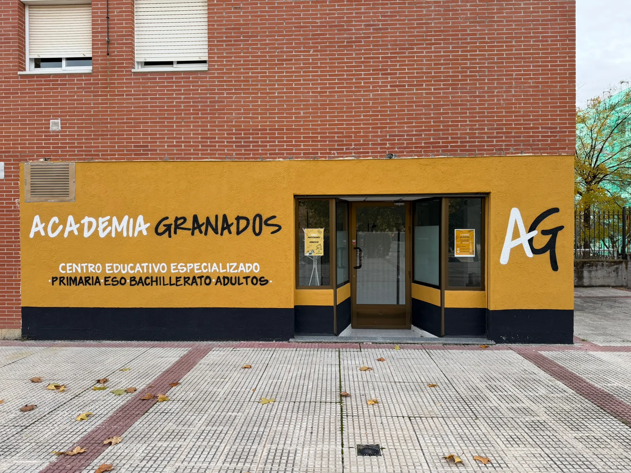 Fachada de Academia Granados: muro amarillo mostaza con el nombre pintado a mano en blanco y negro, y la puerta acristalada de entrada.